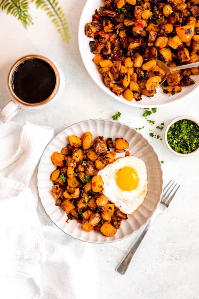 overhead shot of a white breakfast table with a white pasta bowl filled with a hearty portion of baked home fries and a sunny side up egg with a mug of coffee, a pinch bowl of parsley, and the rest of the serving bowl filled with the crispy home fries.