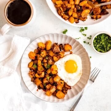 square hero image of a breakfast table set with an earthenware mug filled with blac coffee, a white plate with a serving of baked home fries and a fried egg, a pinch bowl of chopped fresh parsley, and the serving platter with the remaining baked home fries in the top of the frame.