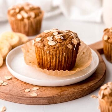 easy, healthy whole wheat banana bread muffins on a white table with the one in the center having the muffin liner peeled off on a white plate.