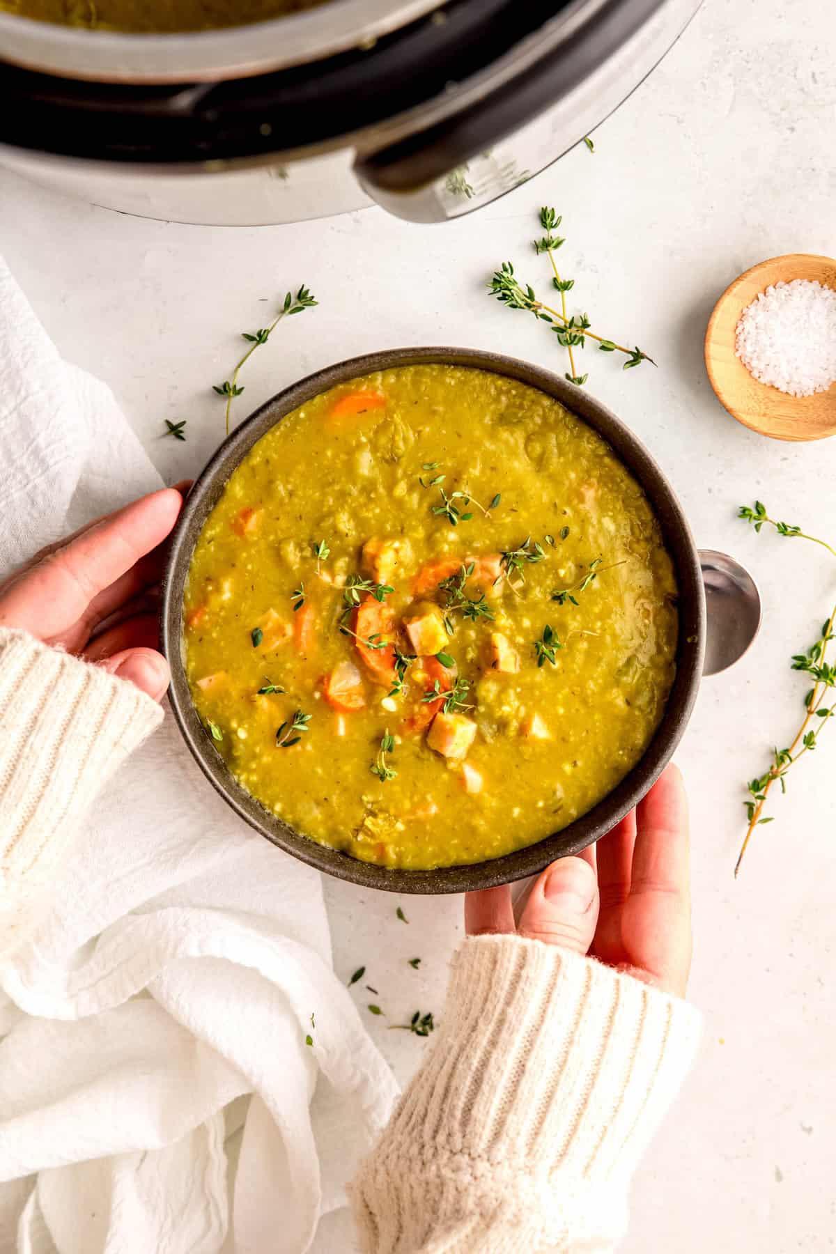 hands in a white sweater up to the knuckles setting a bowl of easy instant pot split pea soup down on a white table.