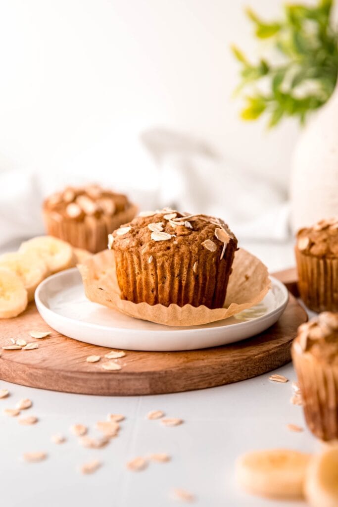 unwrapped healthy whole grain banana muffin on a white plate on a wooden round with more muffins and sliced bananas and rolled oats scattered around.