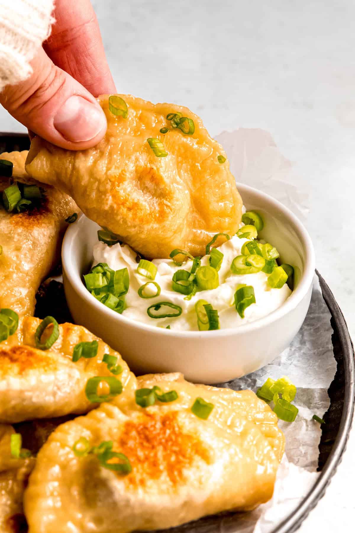 action shot of a hand clad in a white sweater up to the knuckles dipping a pan fried homemade pierogi into sour cream with scallions on top.