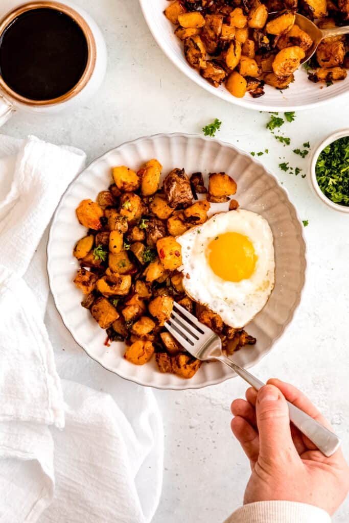 hand holding a silver fork taking a bite of baked home fries with a sunny side up egg and fresh parsley.