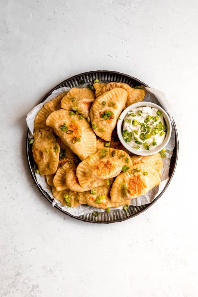 serving platter of potato cheese pierogi after pan frying and adding a bowl of sour cream for dipping and a scattering of scallions for garnish.