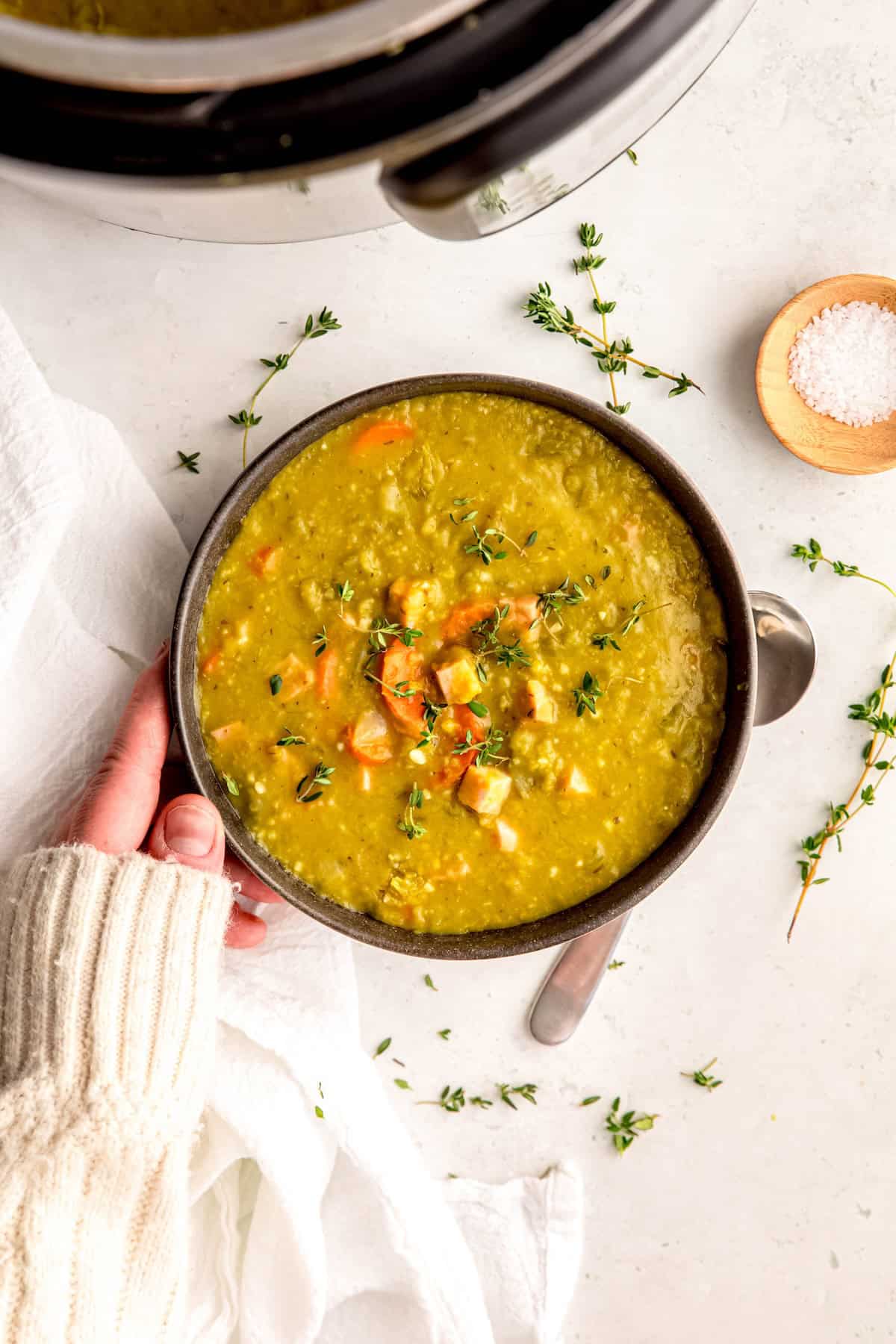 hand clad in a white sweater up to the knuckles adjusting a bowl of instant pot ham and split pea soup on a white table.