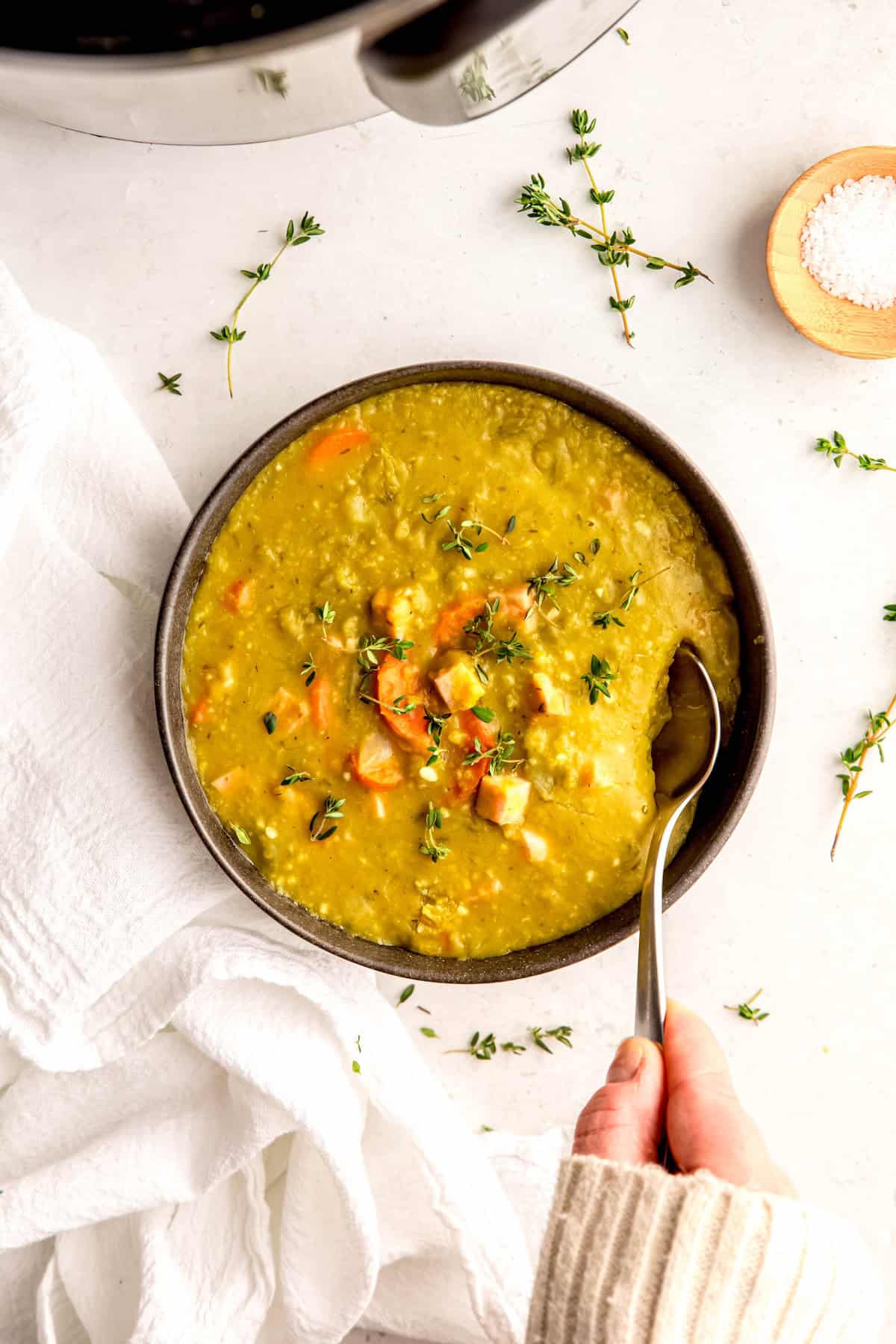 hand clad in white sweater up to the knuckles taking a silver spoonful from a bowl of Instant Pot split pea soup on a white table with fresh sprigs of thyme.