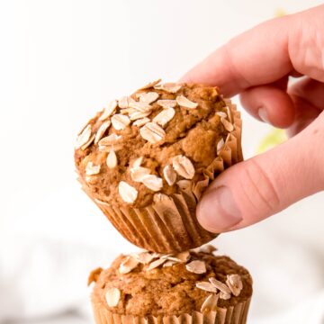 hand taking the top banana bread muffin from a stack of three; the muffin top is tilting towards the frame, showing the oat topping.