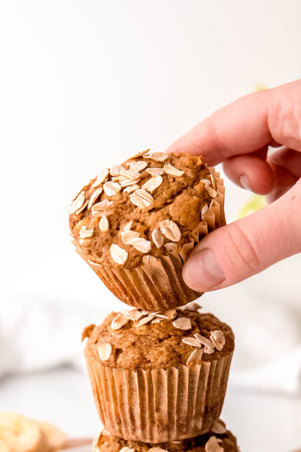 hand taking the top banana bread muffin from a stack of three; the muffin top is tilting towards the frame, showing the oat topping.