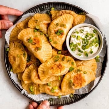 square hero image of a serving platter filled with golden pan fried potato and cheese pierogi with a dipping bowl of sour cream on the side.