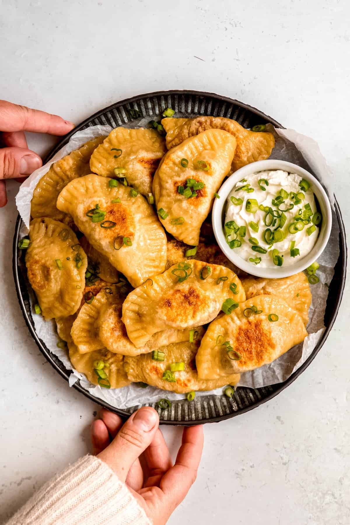 hands setting down a black platter of pan fried potato and cheese pierogis with a small bowl of sour cream with scallions for dipping.