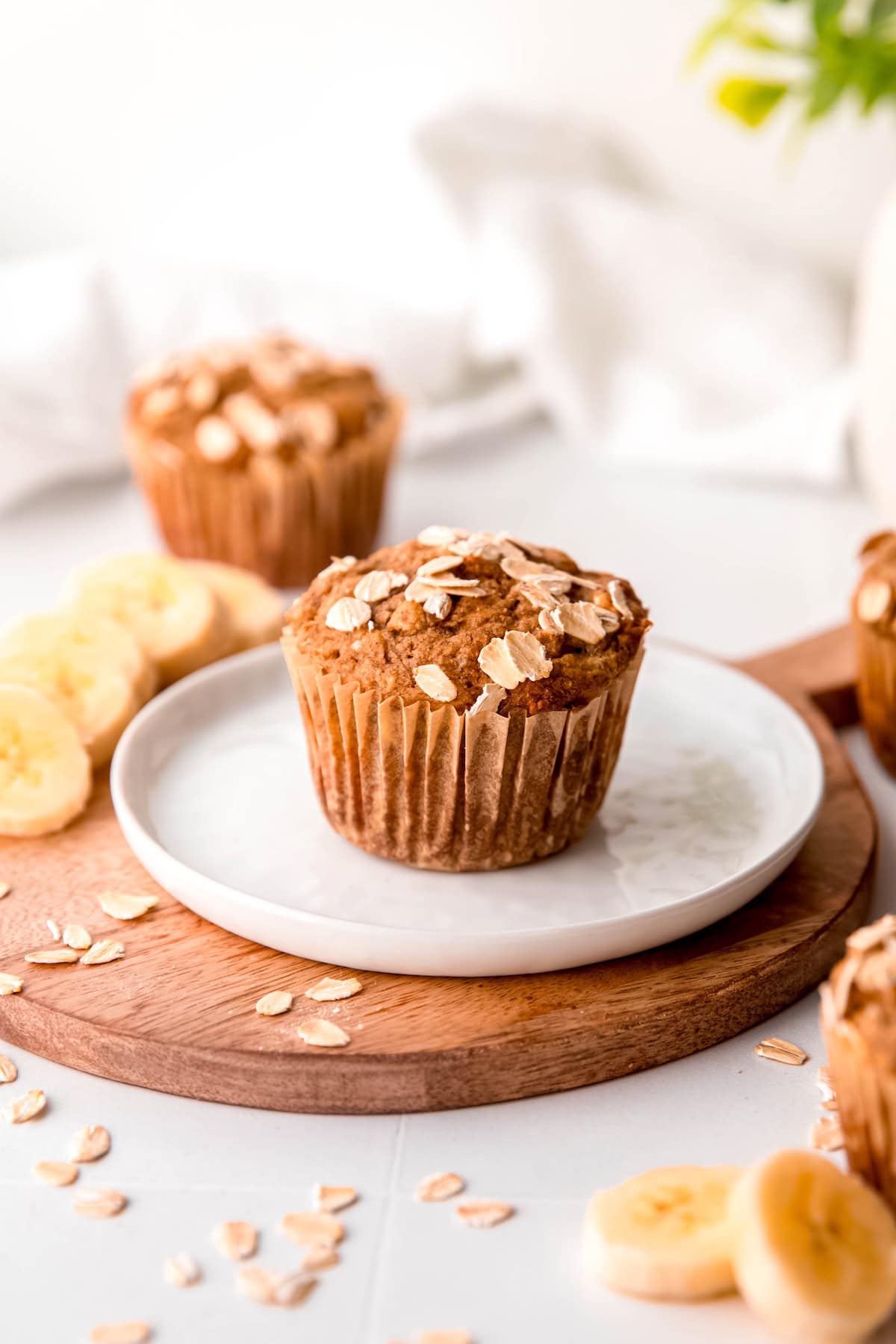 white plate with a whole grain banana bread muffin on a table with fresh banana slices and rolled oats scattered around.