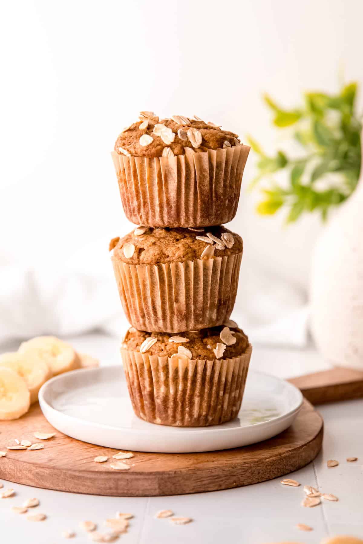 a stack of three healthy whole grain banana bread muffins on a white plate on a wooden round with sliced bananas and rolled oats.