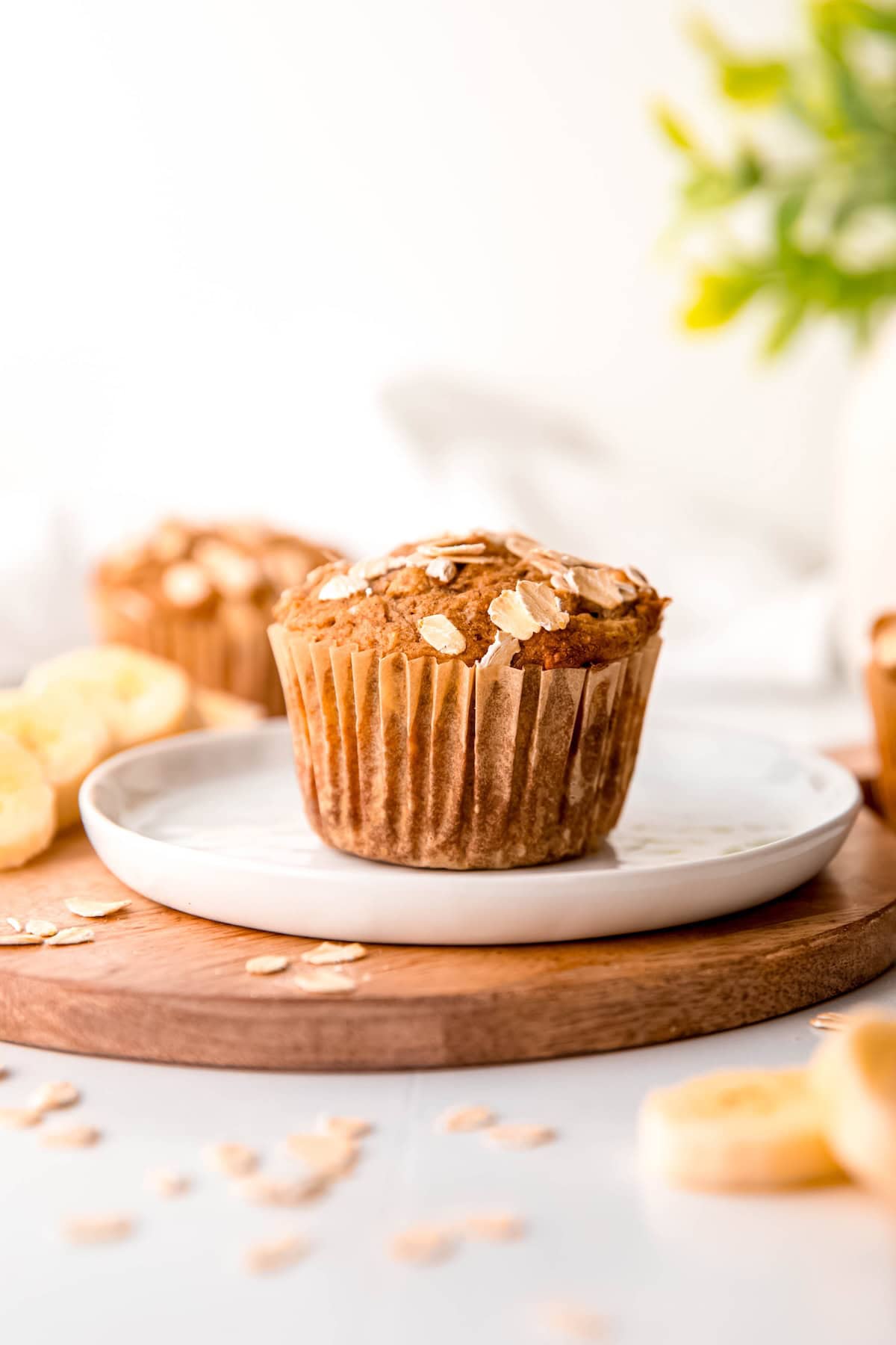 whole wheat banana muffin on a white plate on a wooden round on a white table; more muffins and sliced bananas are blurred out in the background, with banana slices and rolled oats in the foreground.