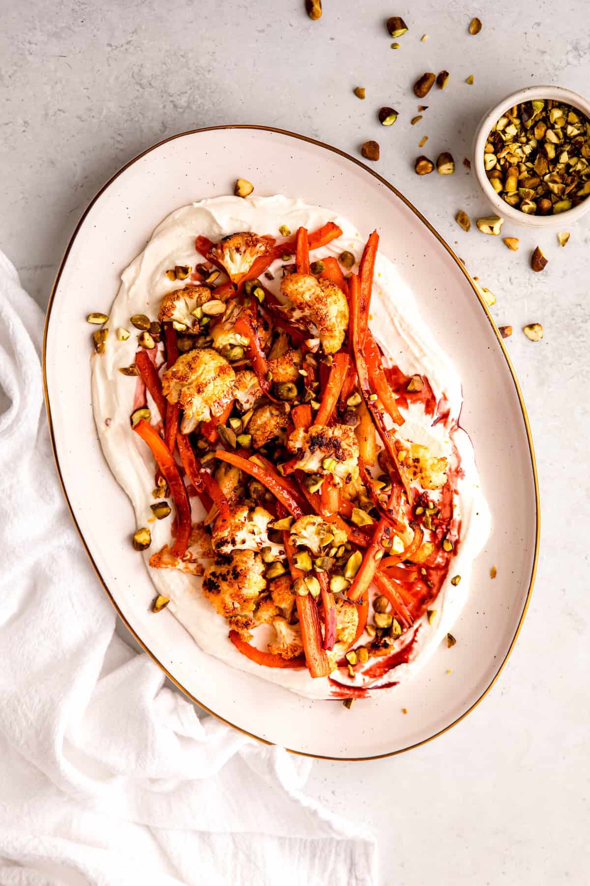 oval white serving platter with whipped feta topped with harissa roasted carrots and cauliflower, a drizzle of pomegranate molasses, and crushed pistachios on a white table with a white pinch bowl with more pistachios.