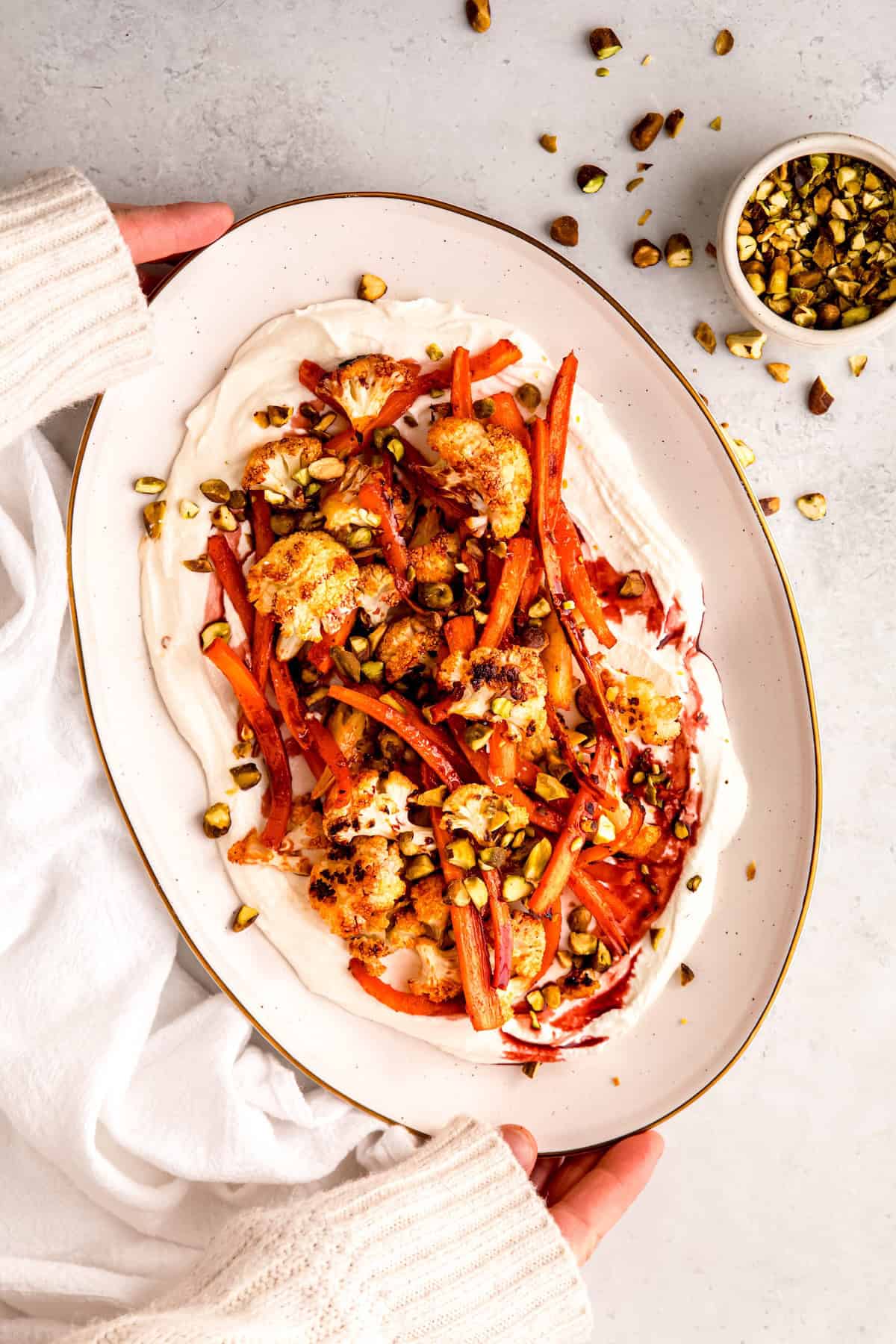 hands clad in a cozy white sweater up to the knuckles setting down a serving platter with spicy mediterranean roasted cauliflower and carrots on a bed of whipped feta onto a white table. .