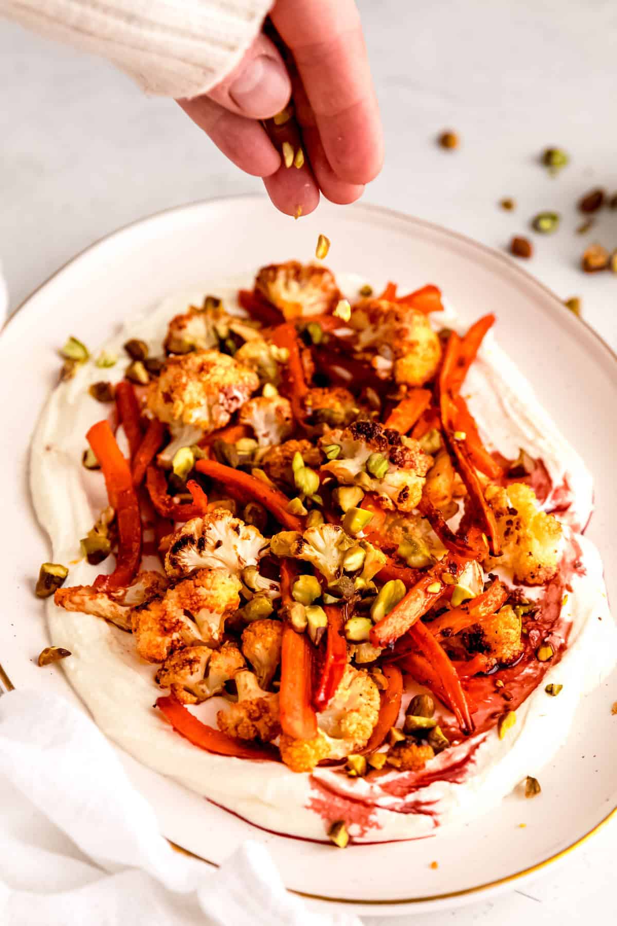 action shot of a hand clad in a white sweater up to the knuckles sprinkling chopped pistachios on top of the spicy roasted carrots and cauliflower.